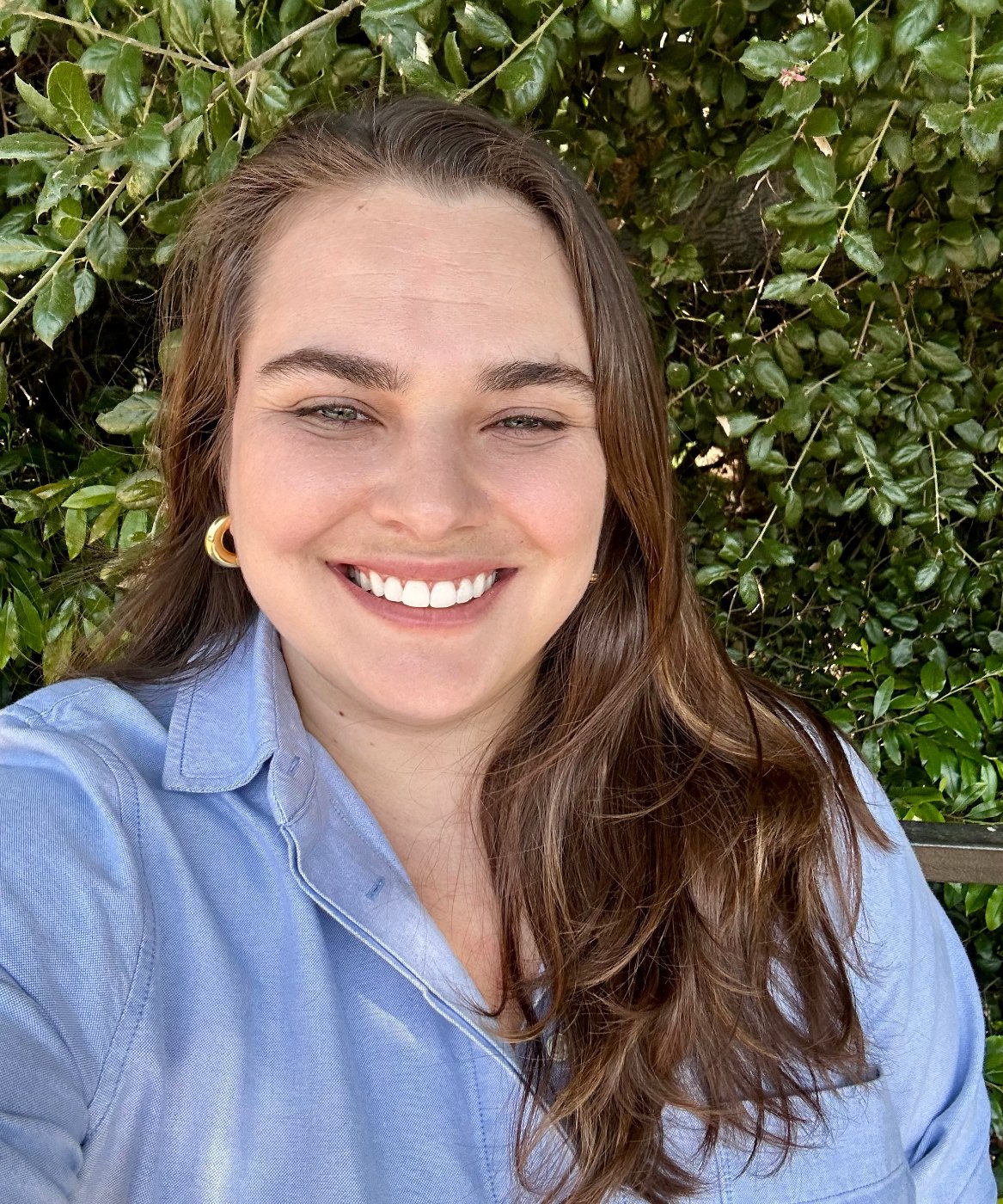 Andrea smiling in a blue button-down shirt in front of green leaves